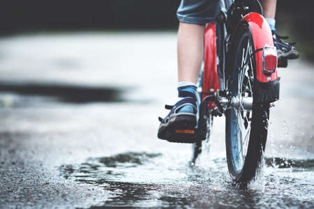 Child on a bicycle at asphalt road in summer. Bike in the park moving through puddle on rainy dayの写真素材