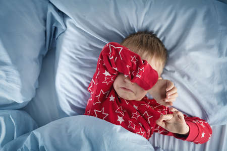Sleepy boy lying in bed with blue beddings. Tired child in bedroom. Little kid lying awake in red pajamas with toothacheの写真素材