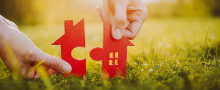 Red house on green grass on a sunny day. Model of home with a shape of two parts of puzzle in human handsの写真素材