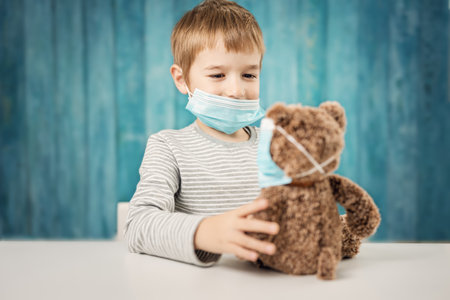 Little boy sitting at the table with a teddy bear in medical facial masksの写真素材