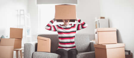 Tired woman with cardboard box on her head sitting on the sofa in her new house.の写真素材