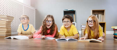 Four cute children in glasses lying on the floor indoors with booksの写真素材