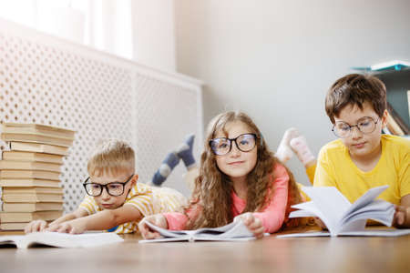 Four cute children in glasses lying on the floor indoors with booksの写真素材