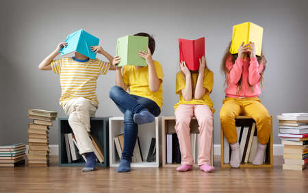 Group of children sitting on the books stacks and on the bookshelves in the room and reading periodical.の写真素材