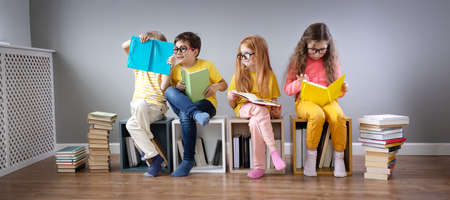 Group of children sitting on the books stacks and on the bookshelves in the room and reading periodical.の写真素材