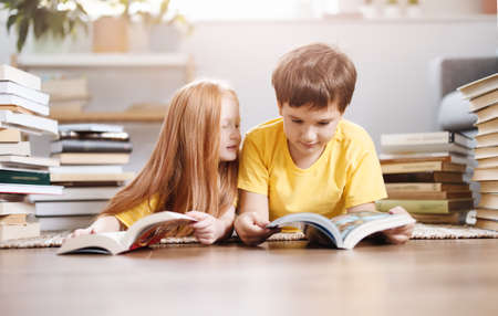 Two cute children lying on the floor indoors with books on the headの写真素材