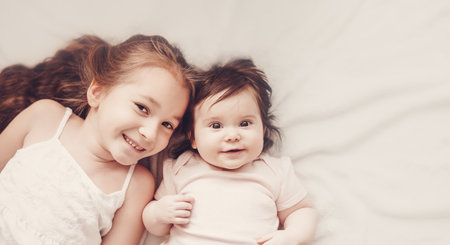 Two smiling sisters lying on the white blanket indoorsの写真素材