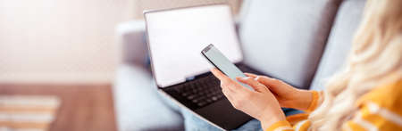 Woman with smartphone in her hands sitting on the couch and working on the laptopの写真素材