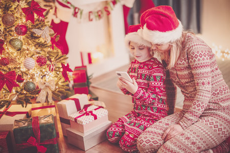 Mother and daughter sitting with smartphone on the floor indoors in Christmas eve.の写真素材