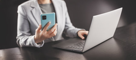 Woman in the office holding in her hands mobile phone and typing on the laptop.の写真素材