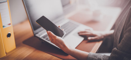 Woman with smartphone in her hand working on the laptop in officeの写真素材