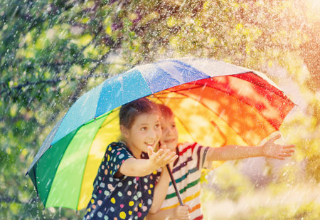 Boy and girl standing outdoors in rainy day under colourful umbrella and trying to catch any raindrops.の写真素材
