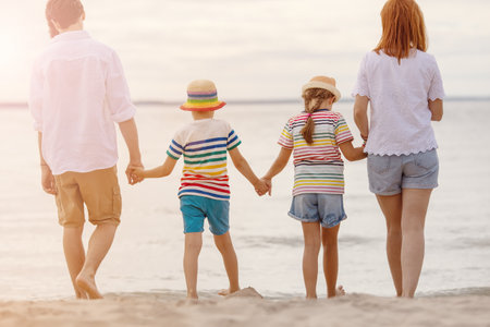A family stands by holding hands on the seashore at the waters edge looking into the distance.の写真素材