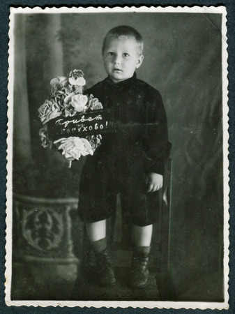USSR - CIRCA 1960s: An antique photo shows little boy standing on a chair and holding flowers with the inscription: "Greetings from Petukhovo"のeditorial素材