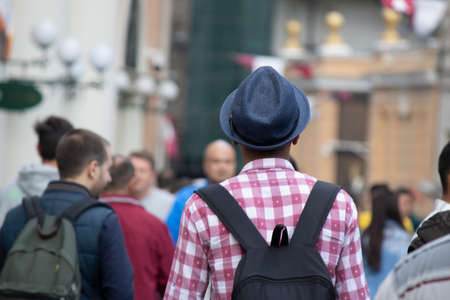 A teenager with a checkered shirt, a fedora and a backpack. The man is turned back and photographed...