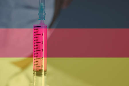 The doctor is holding the syringe with blue liquid in the glove. Close-up. Syringe focused. The doctor has a white outfit. Germany flag in the background.の写真素材
