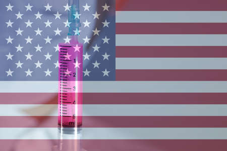 The doctor is holding the syringe with blue liquid in the glove. Close-up. Syringe focused. The doctor has a white outfit. Flag of the United States in the background.の写真素材