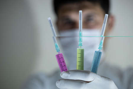 The doctor with a mask in his mouth is holding three vaccines in his hand. Vaccines in red and green colors. Studio shooting. Close up.の写真素材