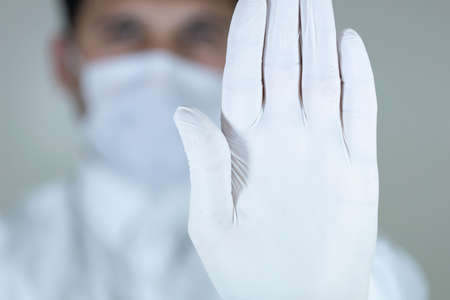 male doctor in a white coat and wihte sterile gloves shows a stop gesture with his right hand,.の写真素材