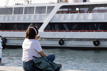 Istanbul, Turkey - November-07.2019: Lady sitting on concrete parquet by the beach. He listens to music with headphones and watches the sea. In front of it is a part of the cruise ship.のeditorial素材