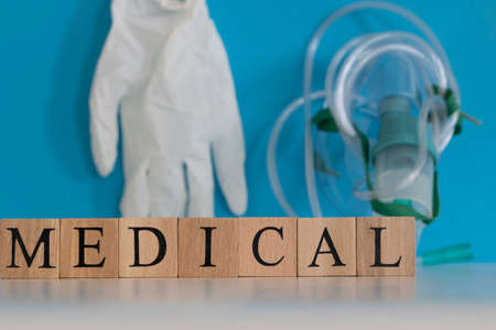Medical text from wooden cubes. White ground and blue. Background oxygen mask and glove objects.