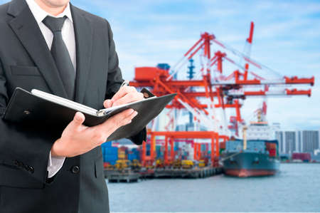 Businessman writing notebook with commercial delivery cargo container ship being unloaded in the...