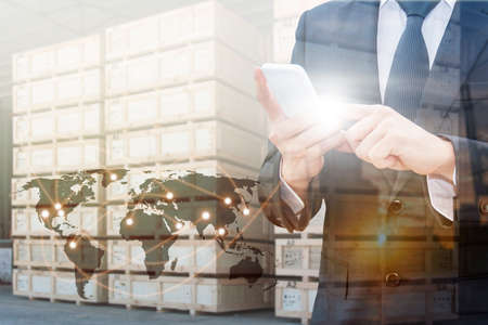 Double exposure of businessman using smart phone network connection world map  , transportation port and blurred cargo warehouse background, international transportation trading business conceptの写真素材