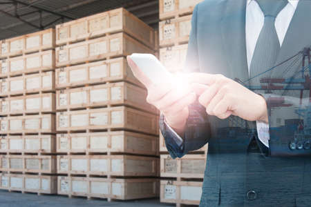 Double exposure of businessman using smart phone and transportation port with blurred cargo warehouse background, international transportation trading business conceptの写真素材