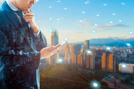 Double exposure of professional businessman connected internet and wireless network with his smart phone and city of business background in business trading and technology conceptの写真素材