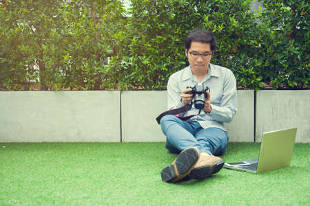 Asian man in casual looking at camera with laptop and smart phone sitting on green grass, relaxing lifestyle or freelance photographer conceptの写真素材