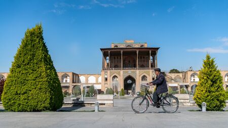 Isfahan, Iran - May 2019: Iranian man riding a bicycle with Aali Qapu Palace in the background in Isfahan Naqsh-e Jahan Square also called Imam Square, Iranのeditorial素材