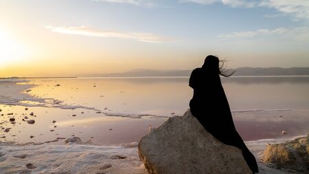 Unidentified Iranian woman in hijab burka sitting on a rock by the Maharloo pink lake in Shiraz, Iranの写真素材