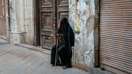 Yazd, Iran - May 2019: Old Iranian muslim woman wearing chador, the black dress, covering her face, sitting by a very old buildingのeditorial素材