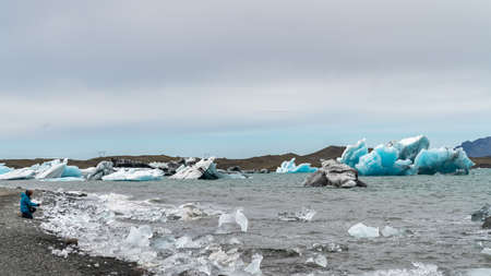 Unidentified woman taking a photo of icebergs in Jokulsarlon glacier lagoon formed with melting ice,...