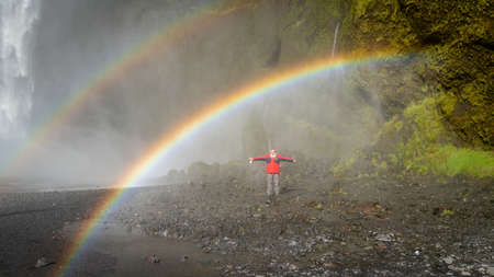 Skogar, Iceland - May 2019: Man standing under the rainbow at Skogafoss waterfall areaのeditorial素材
