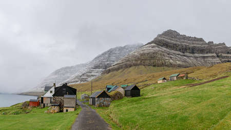 Bour, Faroe Islands - August 2019: A small settlement village overlooking a fjord surrounded by green and snowy mountainsのeditorial素材