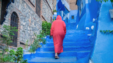 Chefchaouen, Morocco - 11 September 2022: Woman wearing a red burnu walking in the blue roads of Chefchaouen, known as the blue city, famous for its striking blue color painted medina buildingsのeditorial素材