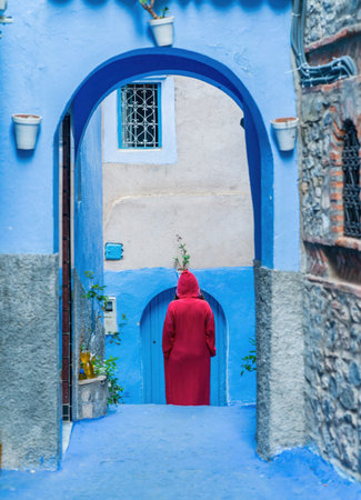 Chefchaouen, Morocco - 11 September 2022: Woman wearing a red burnu walking in the blue roads of Chefchaouen, known as the blue city, famous for its striking blue color painted medina buildingsのeditorial素材