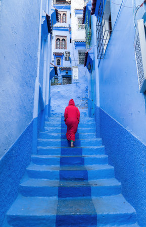 Chefchaouen, Morocco - 11 September 2022: Woman wearing a red burnu walking in the blue roads of Chefchaouen, known as the blue city, famous for its striking blue color painted medina buildingsのeditorial素材