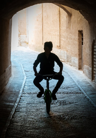 Yazd, Iran - 2 May 2019: Silhouette of a kid riding a bicycle through a dimly lit tunnel in the narrow streets of the ancient city of Yazdの写真素材
