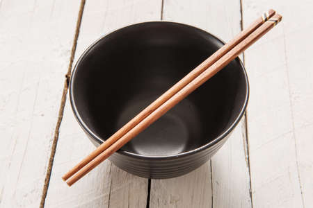 chopsticks lying across an empty bowl that is sitting on a plate on a wooden. The dishes are black.