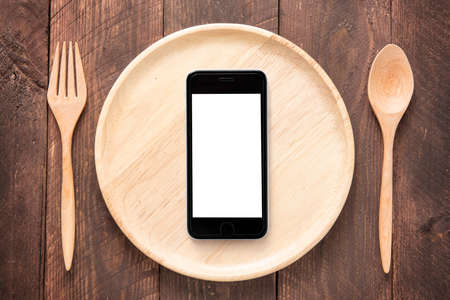 Eating smartphone on set of fork,spoon and dish wood on wooden table.の写真素材