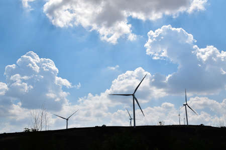 Silhouette of wind turbines on a hills with the background of a blue sky with scattered clouds.