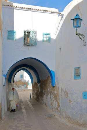 KAIROUAN, TUNISIA - DECEMBER 12, 2019: Typical cobbled and narrow street  inside the historical medina of Kairouan, with blue and white colors, an arcade and a woman traditionally dressed in whiteのeditorial素材