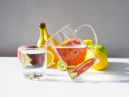 Jug with pure water standing on edge near glass of water and set of fresh fruits on white table. Long shadows on table, space for text above and under objects.の写真素材
