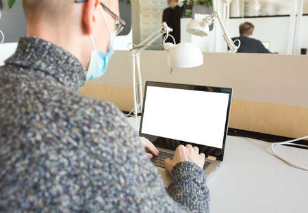 Mockup image of man with face mask working with laptop in office. Business in pandemic time concept.