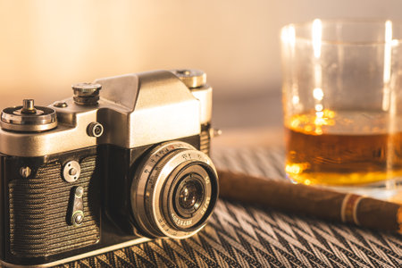 Retro camera paired with a whiskey glass and cigar on a textured table, illuminated by warm, ambient lighting.の写真素材