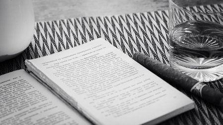 A monochromatic photograph featuring an open book, a cigar, and a glass of water on a textured table...