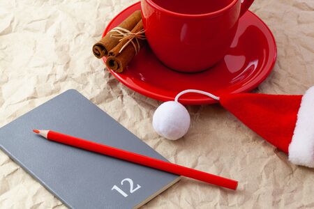 Photo of red cup of coffee, diary with red pencil. Plans for December. Near diary Santa Claus cap. ...