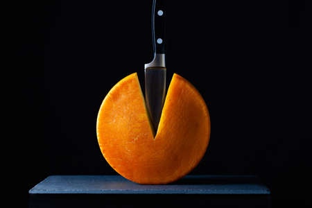 Big round of hard cheese with knife on the slate desk in the dark background.の写真素材